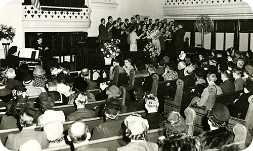 A black and white photo of a crowded church. People are seated in pews, and a choir is standing and singing on stage.