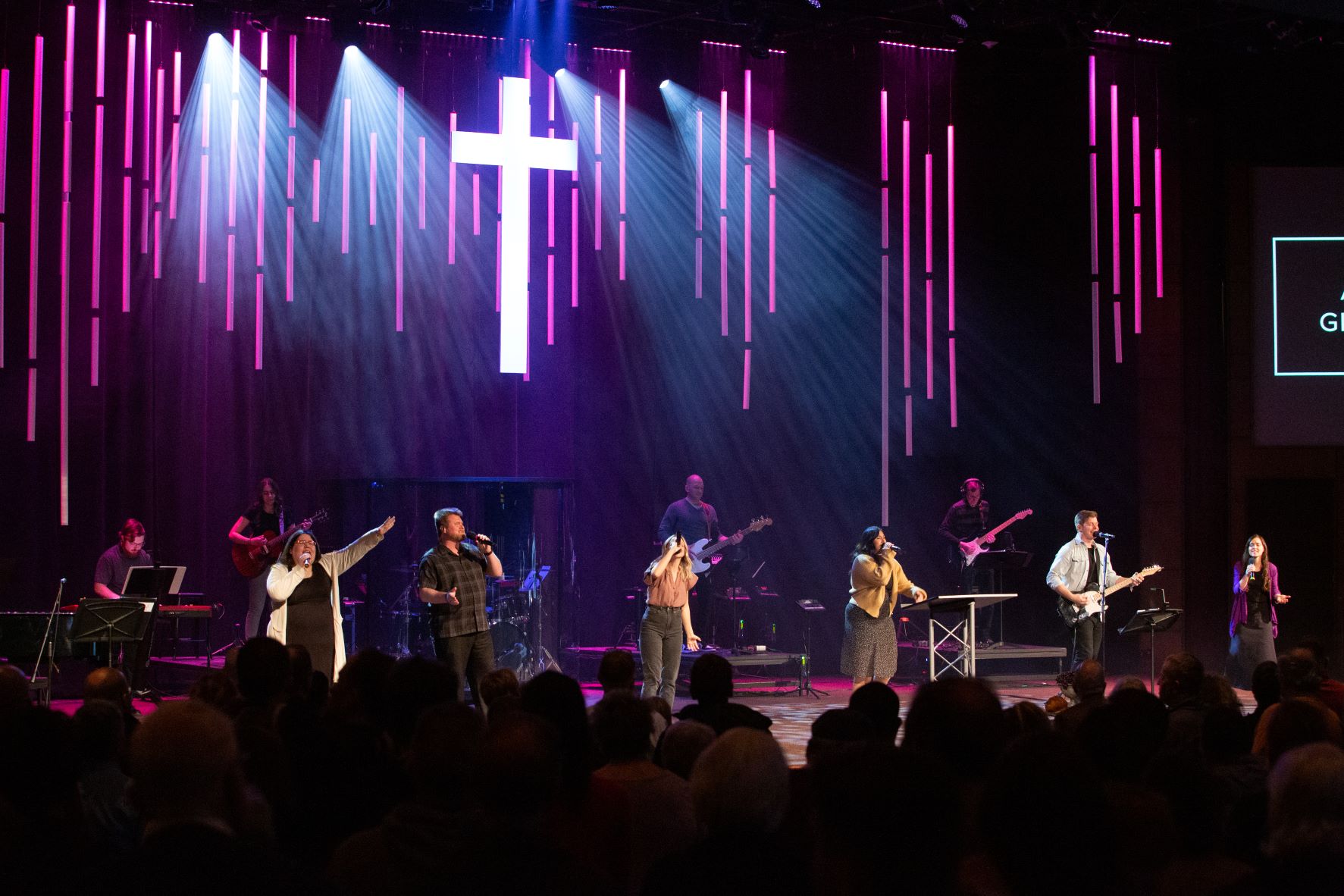  A group of musicians and singers lead worship on a stage illuminated by purple and white lights, with a large glowing cross in the background
