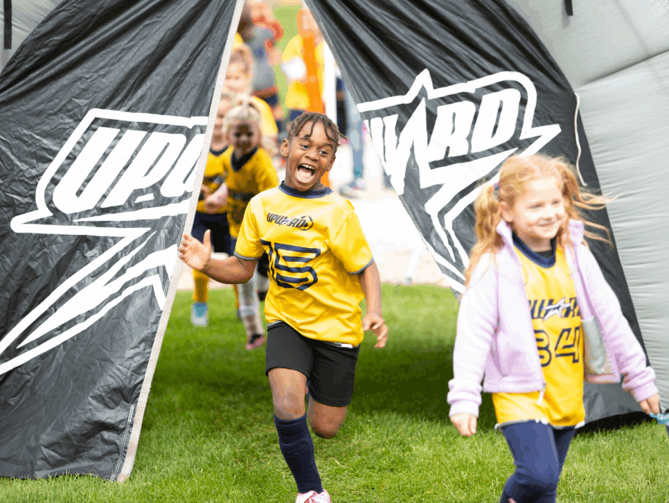 Two children in Upward soccer jerseys run out of the tunnel followed by many more excited players.