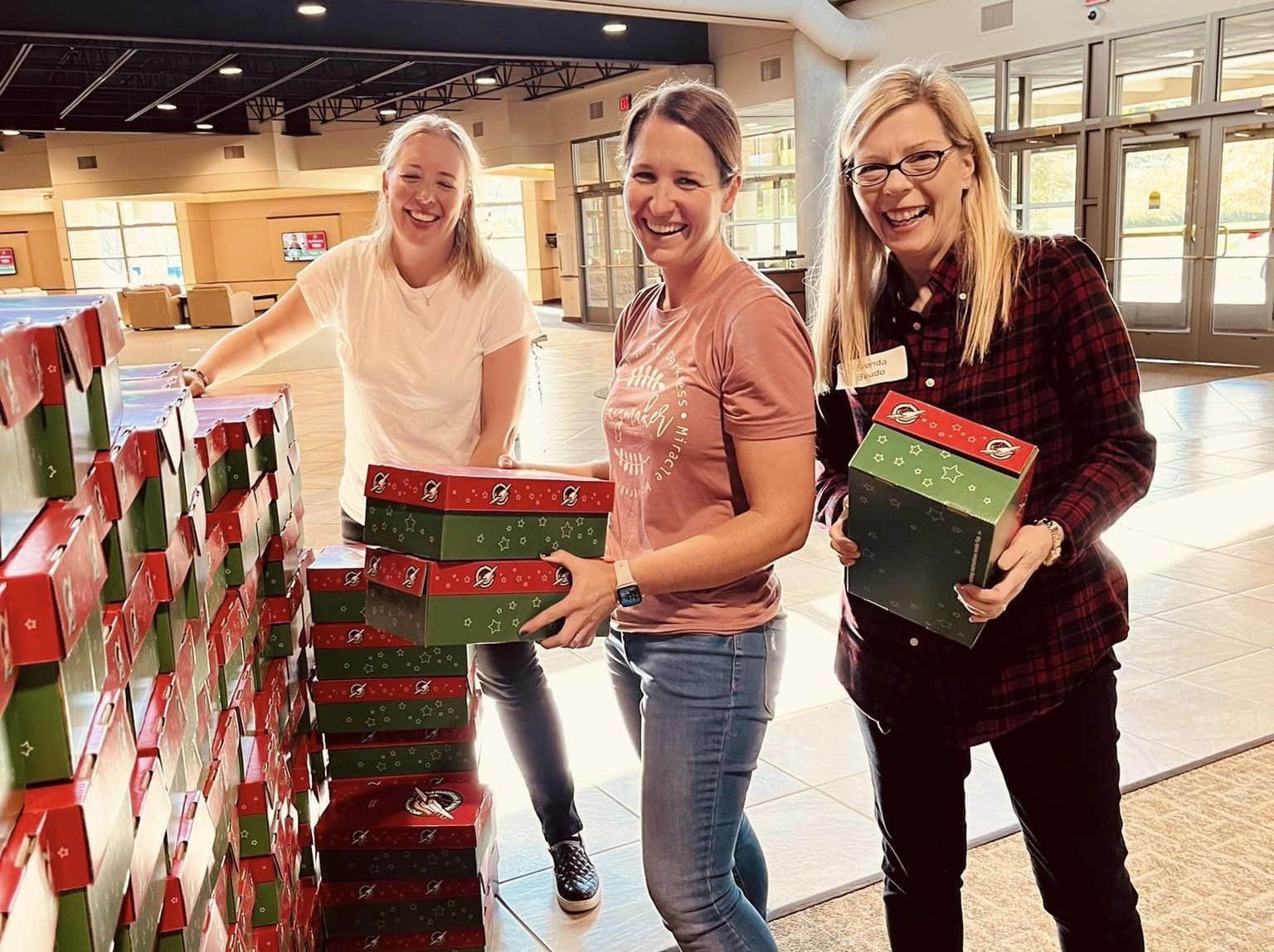 Three women smile as they stack boxes in the foyer of a church.