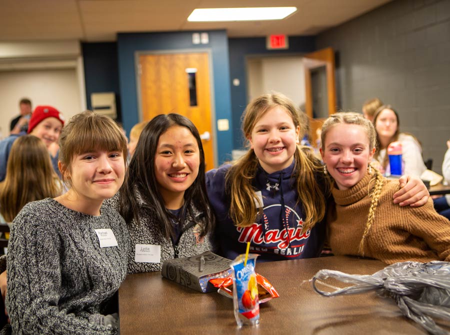 A group of four girls with their arms around each other smile as they enjoy their time in a cafe.