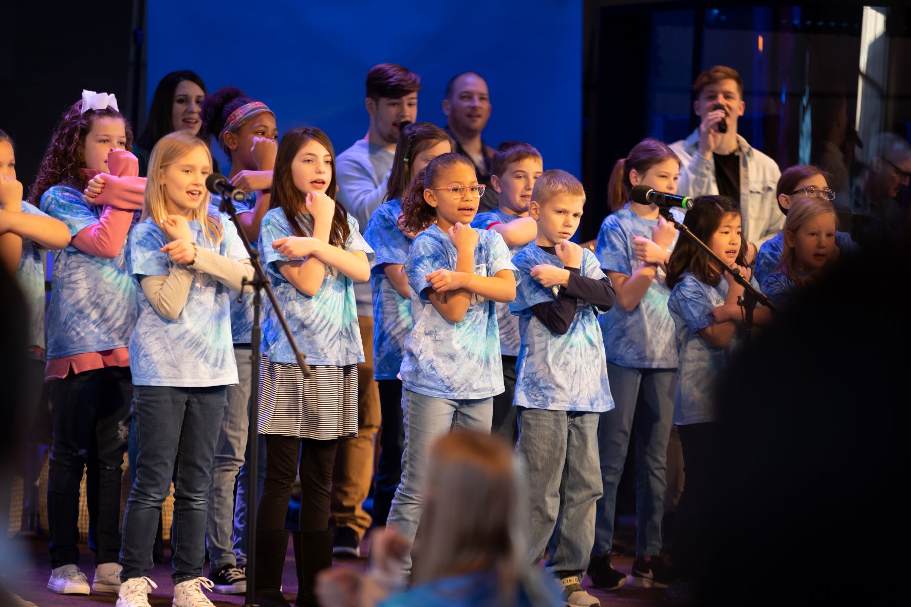 A childrens choir leads worship with choreography accompanied by a band in the background.